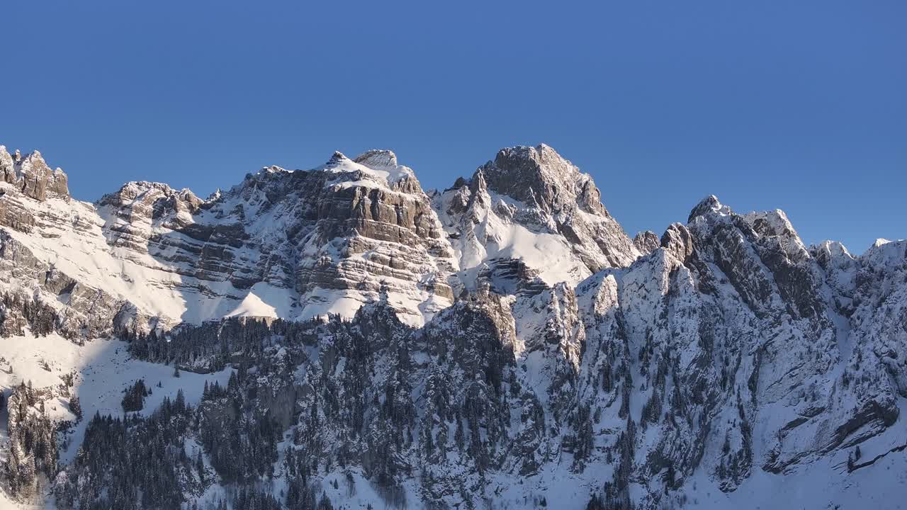 oaring aerial view showcases the majestic Fronalpstock mountain range in Glarus, Switzerland. Snow-capped peaks pierce a brilliant blue sky, creating a breathtaking winter landscape.