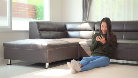 A-cute-young-woman-sitting-on-the-floor-enjoys-interacting-with-her-smartphone