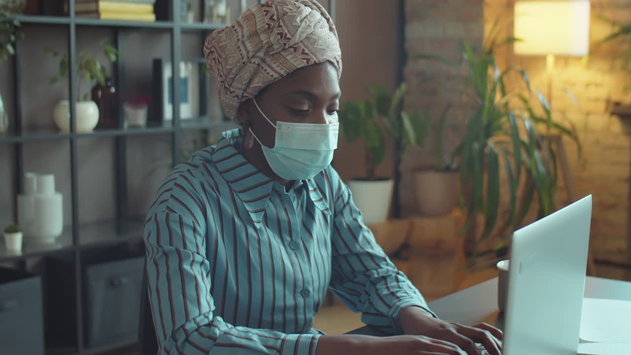 черная бизнесменка в маске работает на ноутбуке в офисе