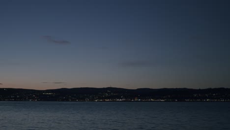 twilight view of a calm body of water with city lights twinkling in the distance under a clear, darkening sky