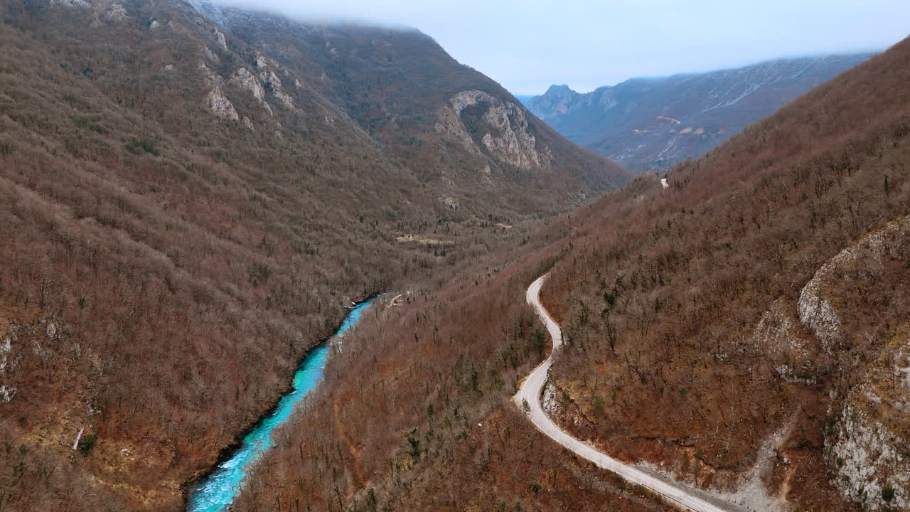 Winding mountain road following a turquoise river in the valley. Aerial landscape of an asphalt road and blue river winding through mountains with leafless winter trees