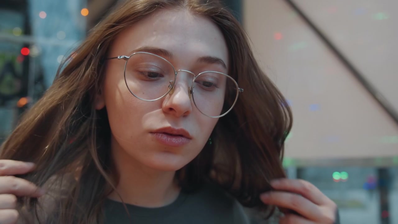Donna che si sistema gli occhiali, ritratto pensieroso in interni sotto luci colorate, dita che toccano capelli e fronte, primo piano intimo con finestra riflettente, momento di pendolare urbano e tranquilla introspezione.
