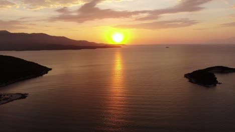 Drohnenansicht-In-Albanien,-Die-Bei-Sonnenuntergang-über-Blauem-Kristallklarem-Wasser-Fliegt,-Wobei-Die-Sonne-Am-Horizont-Mit-Kleinen-Inseln-In-Ksamil-Untergeht