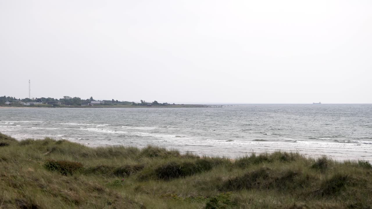 Super slow-motion shot of a Swedish beach on an overcast day. Grass sways in the foreground, while in the distance, a peninsula with traces of civilization emerges under clouds, rain is imminent.