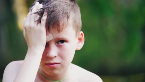 unhappy boy with closed wound on face outdoors. young teenager has a pain from his injury on eyebrow. sad child touches his painful wound in a summertime.
