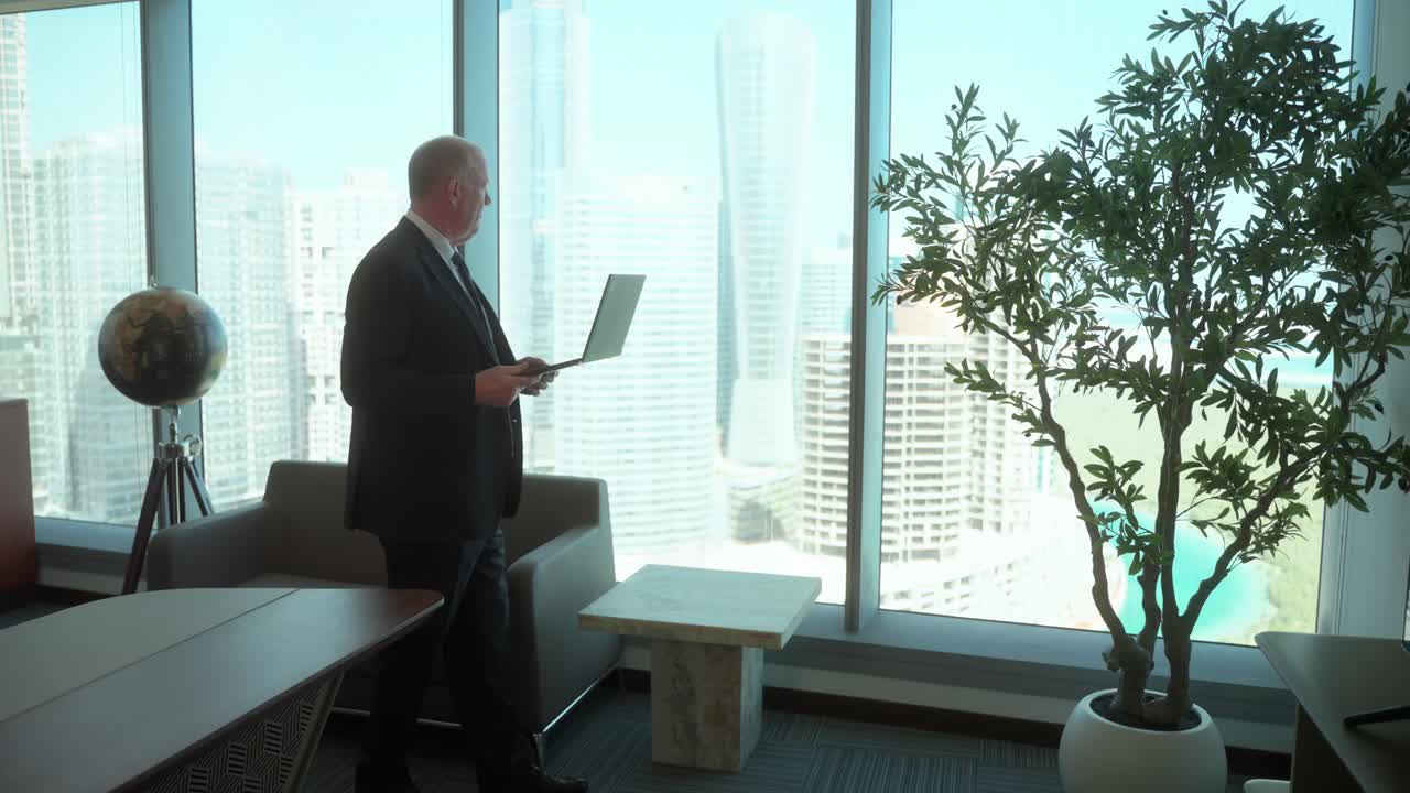 Senior man in a skyscraper office pacing with a laptop in hand while on a web call or considering a business plan