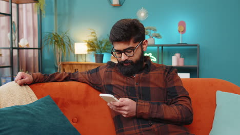 Happy-Indian-man-thinking-texting-messaging-on-smartphone-internet-social-media-app-on-home-sofa