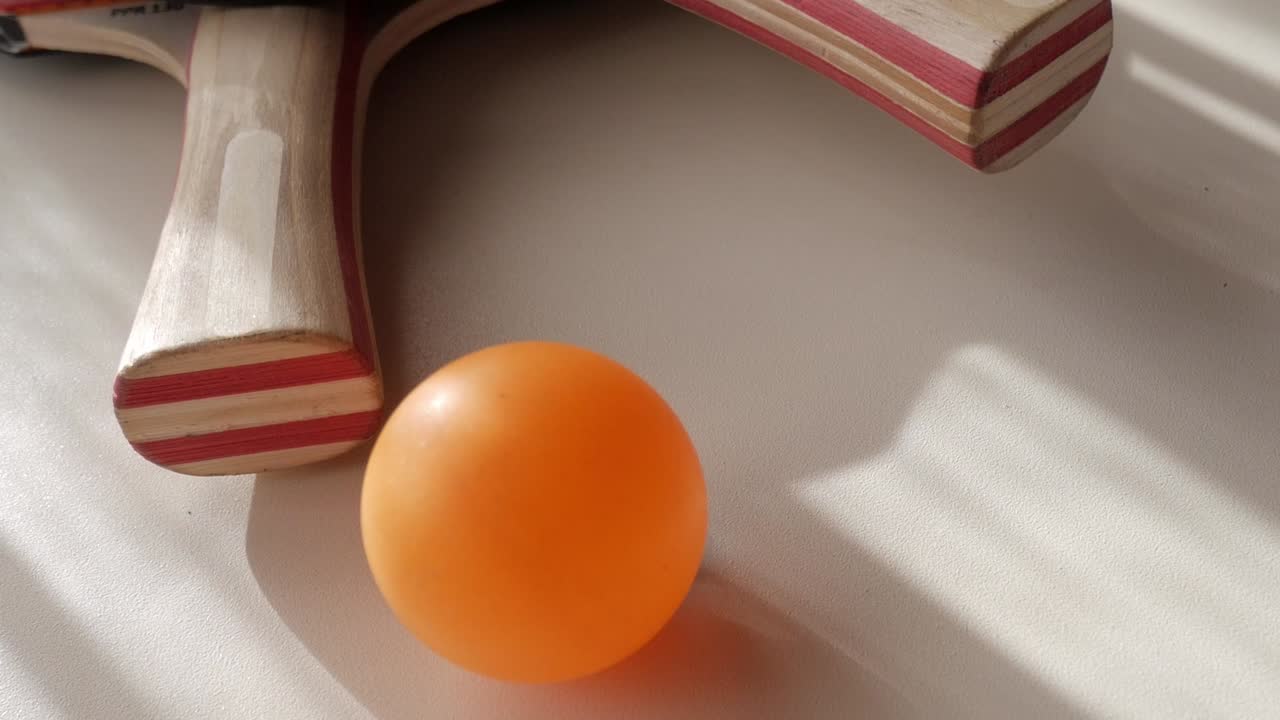 Orange ping pong ball resting near a paddle on a light-colored surface, showcasing a close-up shot of the equipment in a serene setting