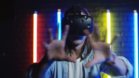 close up view of young man in the vr glasses moving his hands in the air in a room with colorful neon lamps on the wall 1