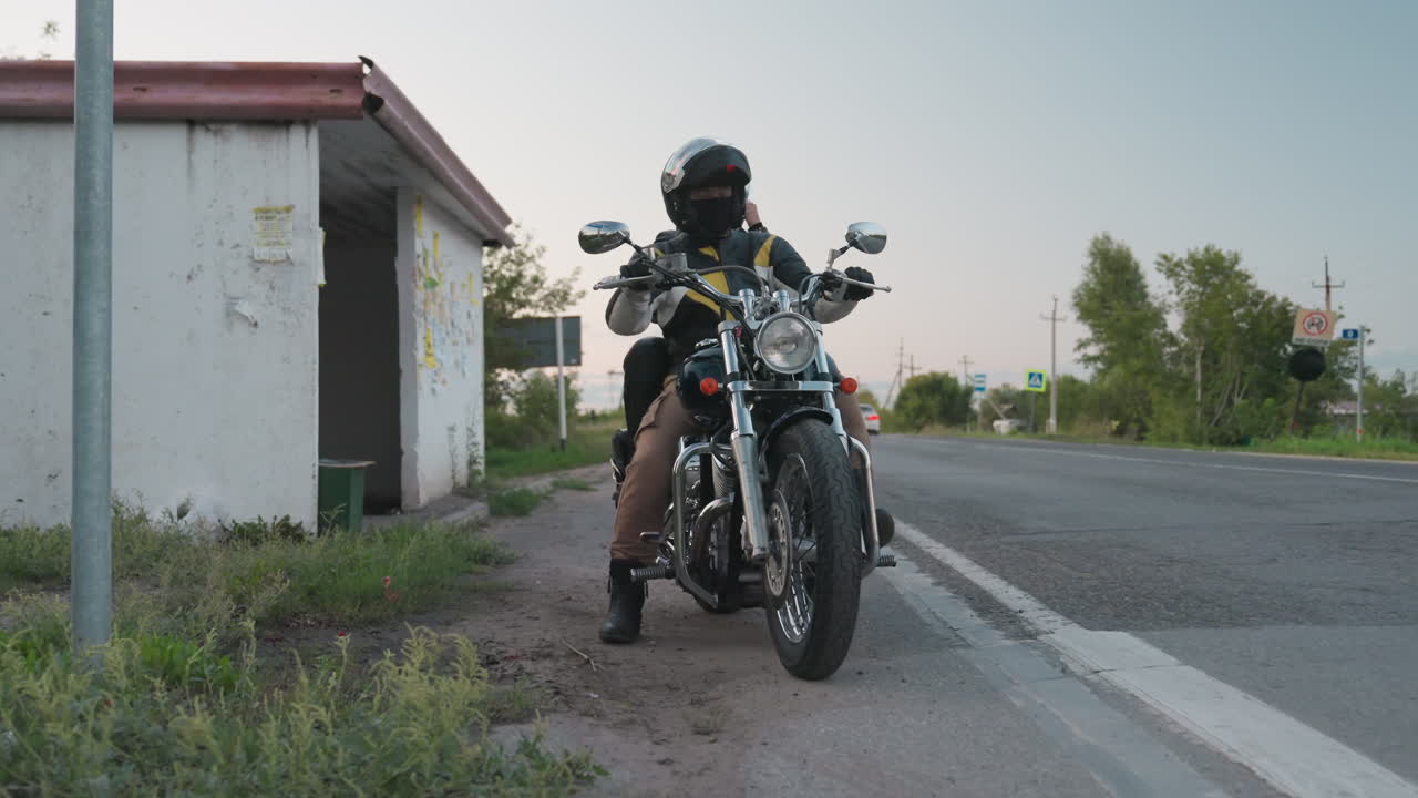 Motorbike rider in helmet starts engine as lights turn on, sitting near roadside beside worn bus stop structure, passenger seated behind, chrome motorcycle reflecting evening glow