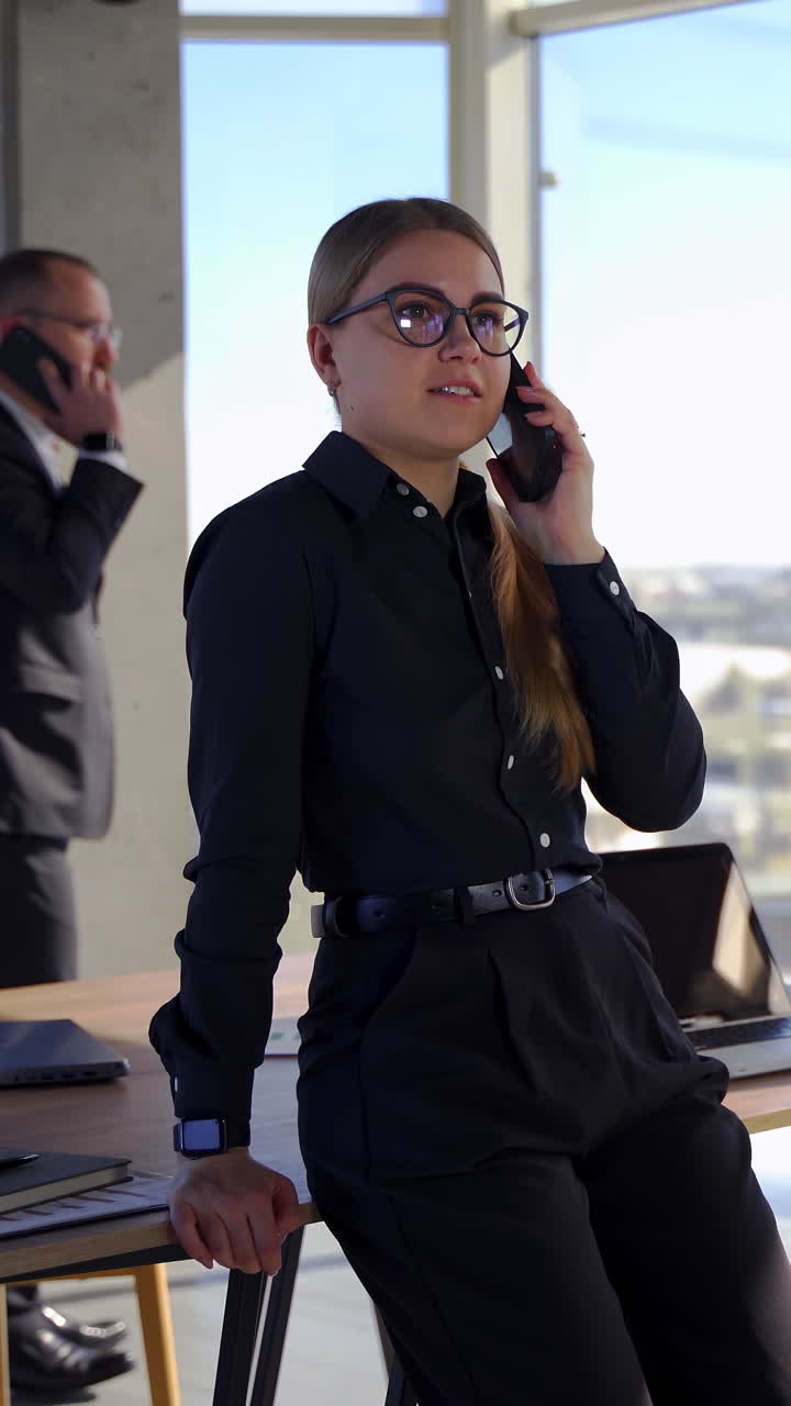 Beautiful secretary and her boss in office. Smiling young business woman holding a phone on the background of a businessman and talking the phone. Vertical video