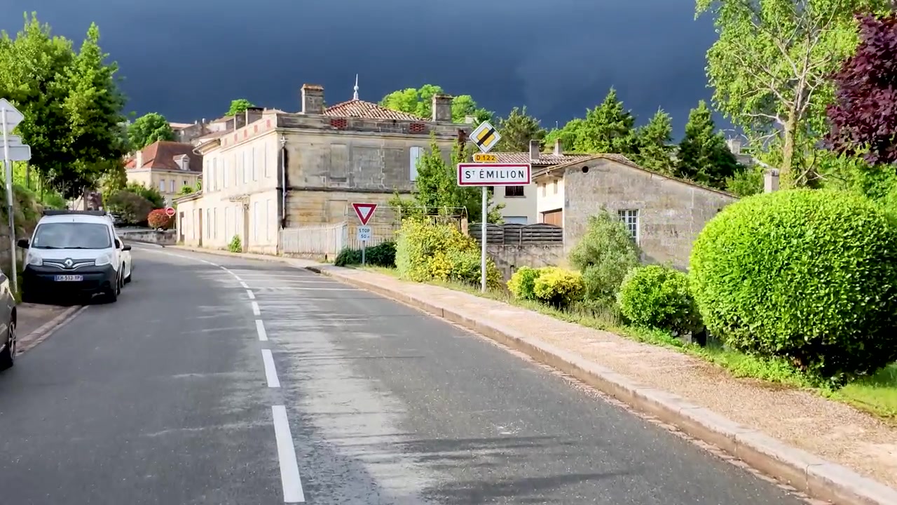 Una telecamera ad angolo basso avanza lungo una strada verso edifici in pietra sotto drammatiche nuvole temporalesche