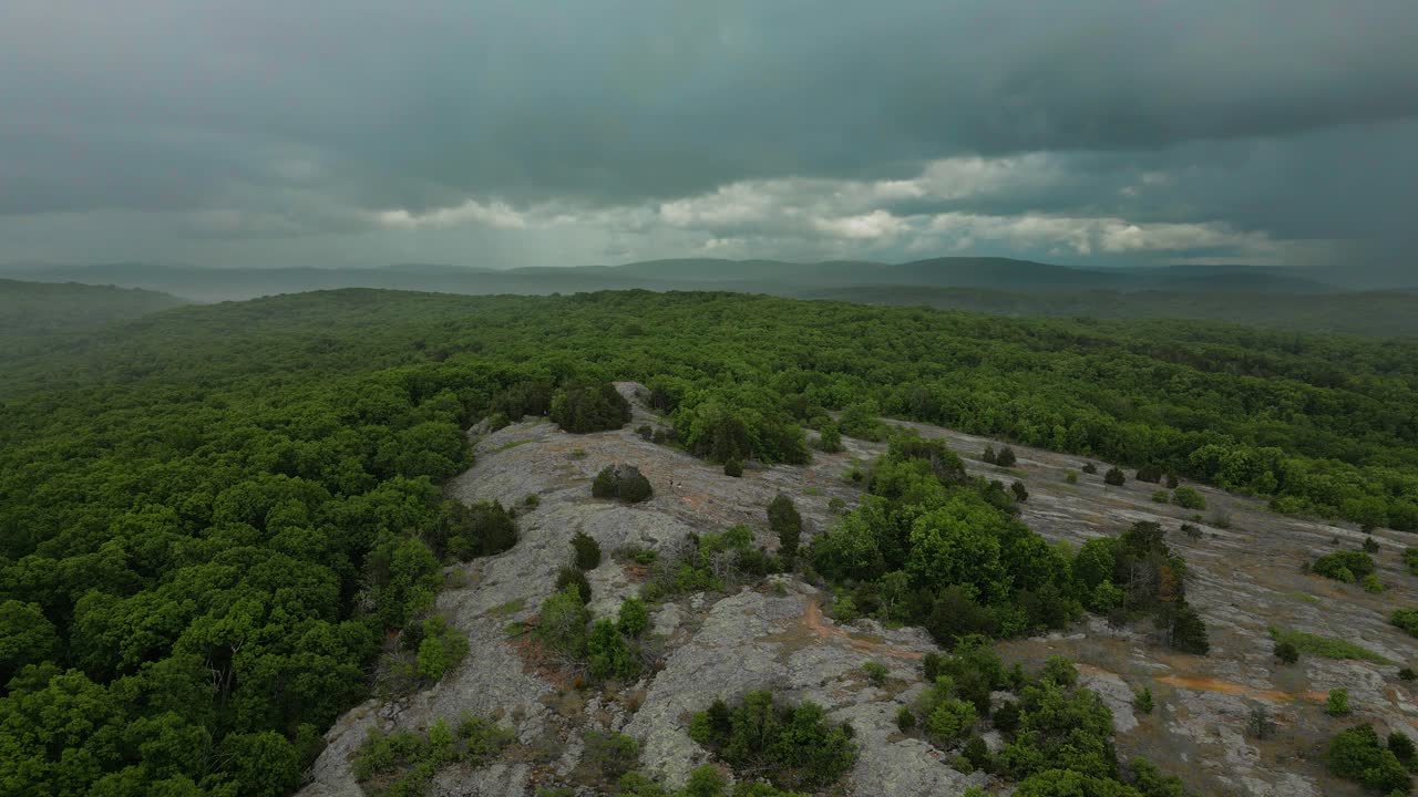 A shot orbiting around the peak of Hughe's Mountain in Missouri while hikers climb down the slope. Thunderstorms can be seen rolling in over the Ozark Mountains in the distance.