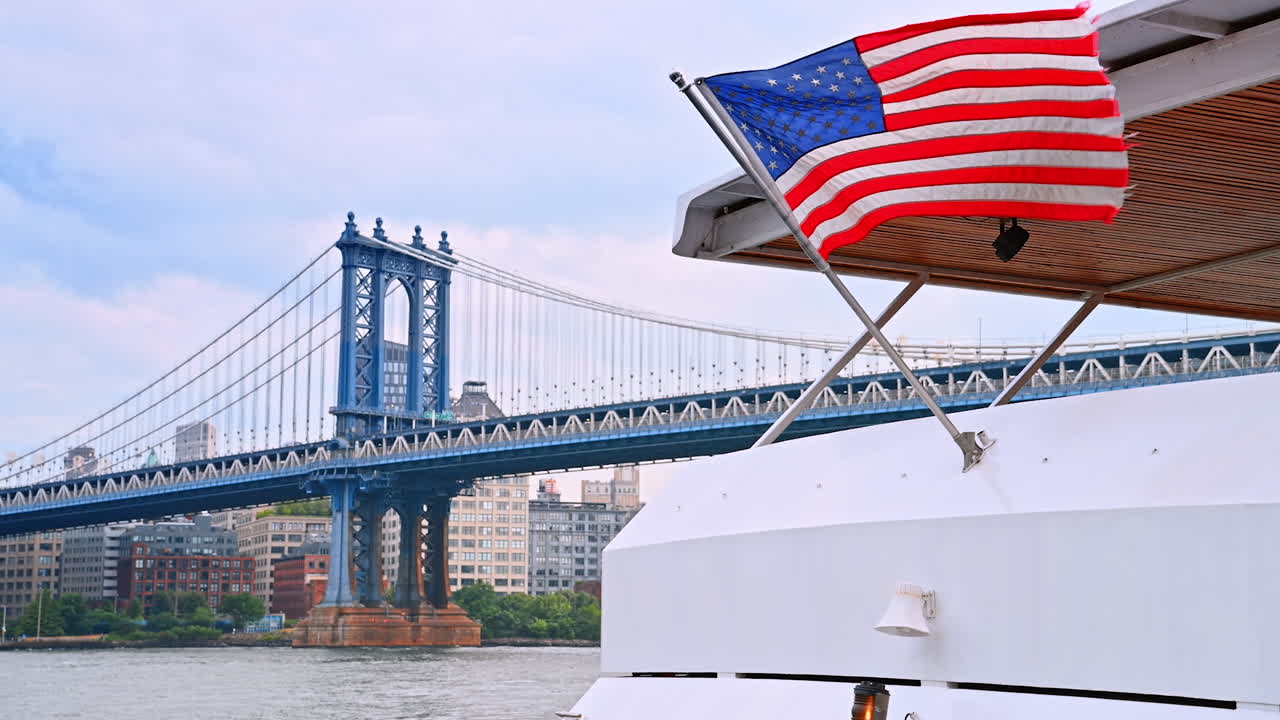 Vista del Manhattan Bridge da una barca. Una barca naviga tranquillamente sotto il Manhattan Bridge, mostrando la bandiera americana e lo skyline della città al crepuscolo
