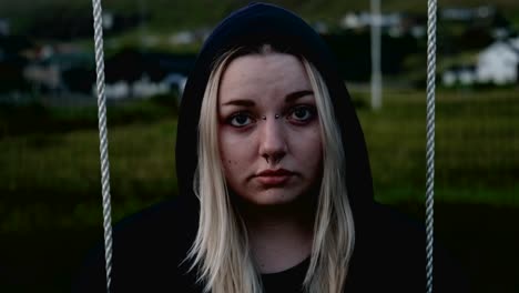Young-blonde-woman-sitting-on-swing-wearing-a-hoodie-looking-sad-into-lens-on-a-cold-spring-evening