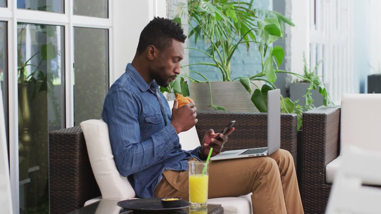 Uomo d'affari afroamericano che usa il laptop, lo smartphone e mangia un croissant in un bar