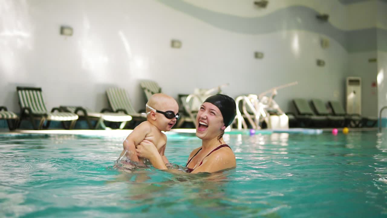 젊은 어머니는 보호 안경을 입은 작은 아들을 들고 수영장에서 수영하는 법을 가르치고 있습니다. 아기가 그의 발로  ⁇ 고