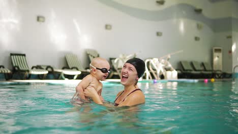 madre joven sosteniendo a su hijo pequeño en gafas protectoras y enseñando a nadar en la piscina. bebé pateando con su