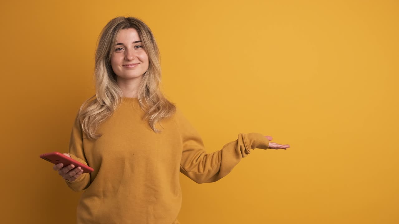 Cheerful young woman with smartphone showing empty wall space on yellow background