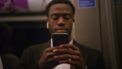 close-up de um jovem empresário de terno que se desloca para o trabalho sentado no metrô de londres usando fones de ouvido sem fio para transmitir de um telefone móvel filmado em tempo real 1