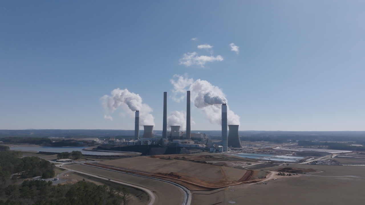 Aerial video rotating around a large power plant with multiple smokestacks and cooling towers releasing steam into a clear blue sky in Georgia.