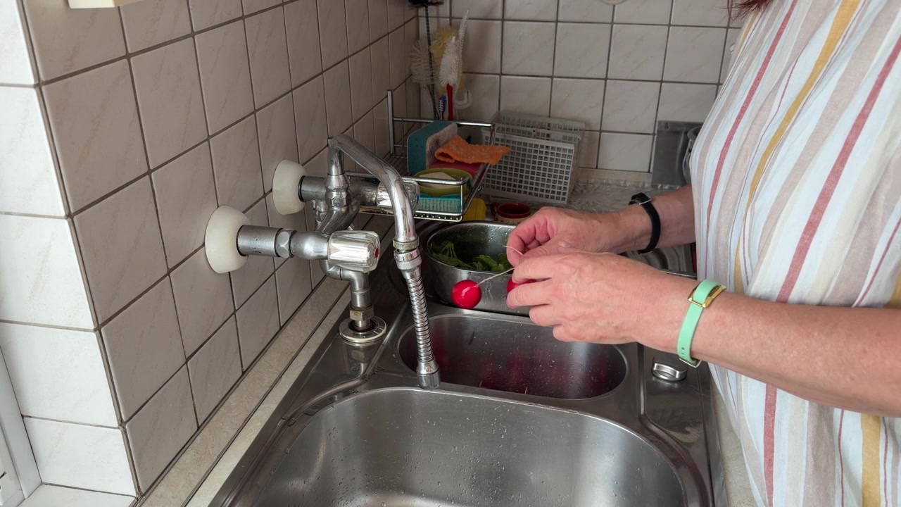 Nærbilde av en ugjenkjennelig kvinne som kutter de tynne røttene fra nyvaskede røde reddiker ved en kjøkkenvask, med rene reddikblader liggende i nærheten under matlagingen, statisk bilde