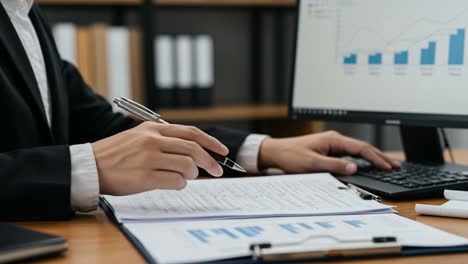 a professional in business attire analyzing data on a computer while taking notes in an office setting, demonstrating concentration and productivity
