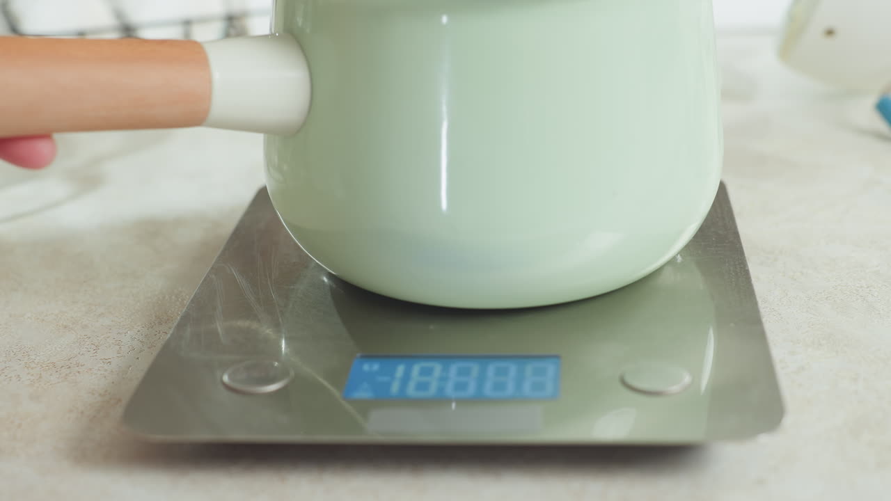 Close up of modern mint green pot with wooden handle being carefully placed on digital kitchen scale during cooking prep in bright kitchen, stainless surface and scale display clearly visible