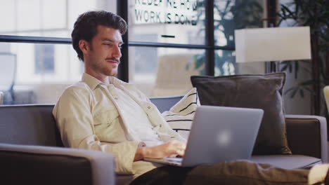 Smiling-young-man-working-on-a-laptop-sitting-with-his-feet-up-in-an-office-lounge-area,-close-up