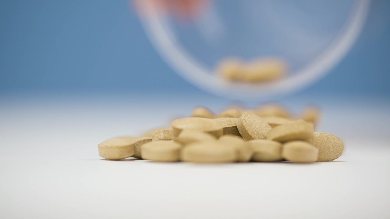 Pillole mediche che si rovesciano da un contenitore di plastica trasparente su una superficie bianca, sfondo blu sfocato.