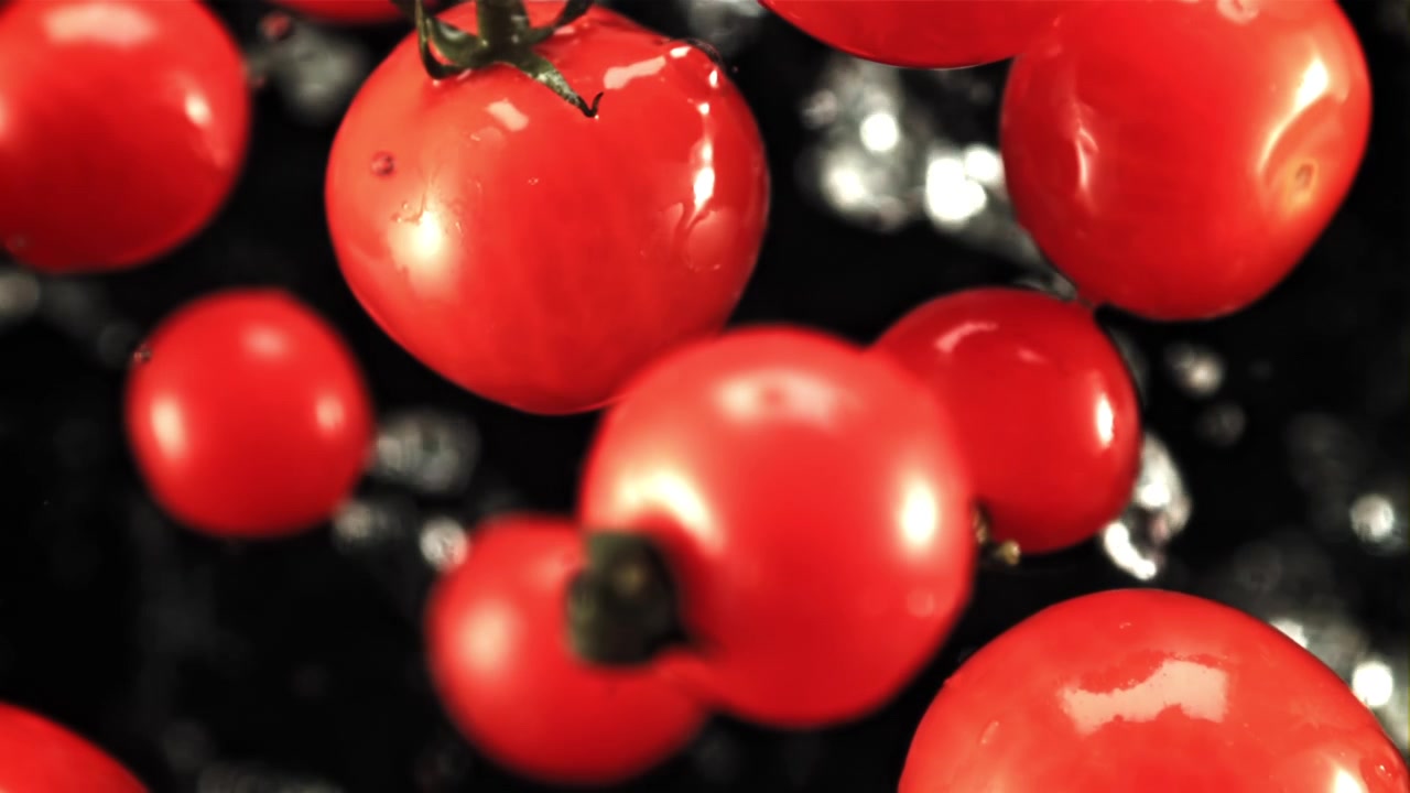 Wassertropfen spritzen auf rote Kirschtomaten vor schwarzem Hintergrund.