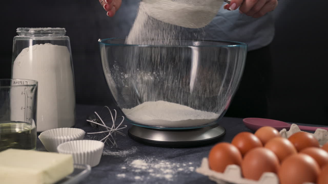 Close up of flour measured sifting into a bowl as the first step in cupcake preparation with eggs and other materials on table, slow motion