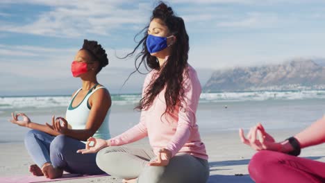 group of diverse female friends wearing face masks meditating at the beach