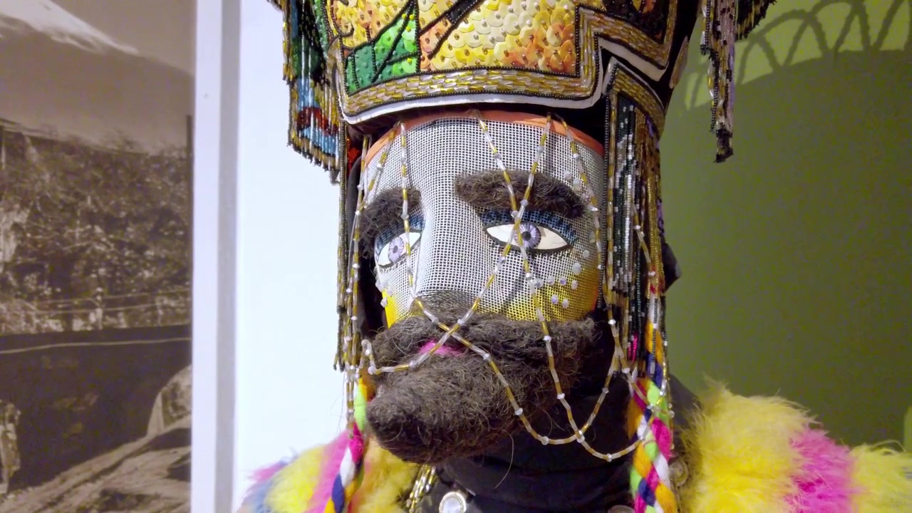 Nærbillede af traditionelt maskeret kostume med broderet hovedbeklædning, malet ansigt og dekorativt skæg udstillet i en kulturhistorisk museumsudstilling i Mexico.