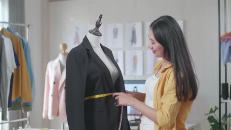 smiling asian female designer measuring business jacket on a mannequin while designing clothes in the studio