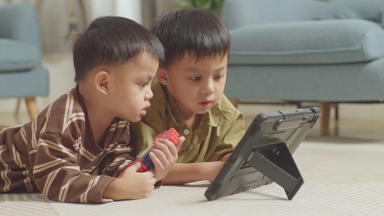 플라스틱 장난감 벽돌을 가진 아시아 어린이들이 바닥에 누워서 집에서 함께 태블릿에서 영화를 보고 있습니다.