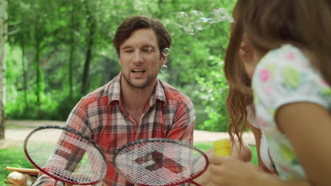 Woman-and-man-playing-with-children-in-park.-Girl-and-boy-blowing-soap-bubbles