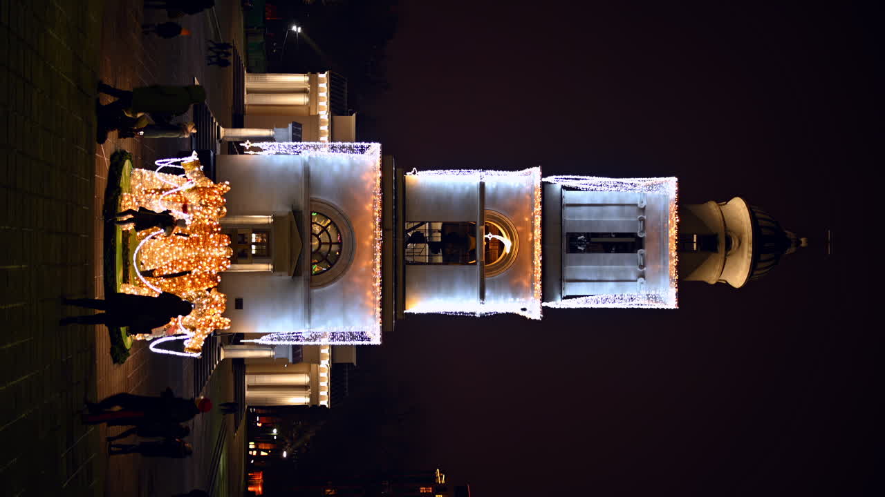 Chisinau, Moldova - December 20, 2021: A historic clock tower shines brightly with festive lights at night. People gather around, enjoying the holiday atmosphere and celebrating together.. Vertical