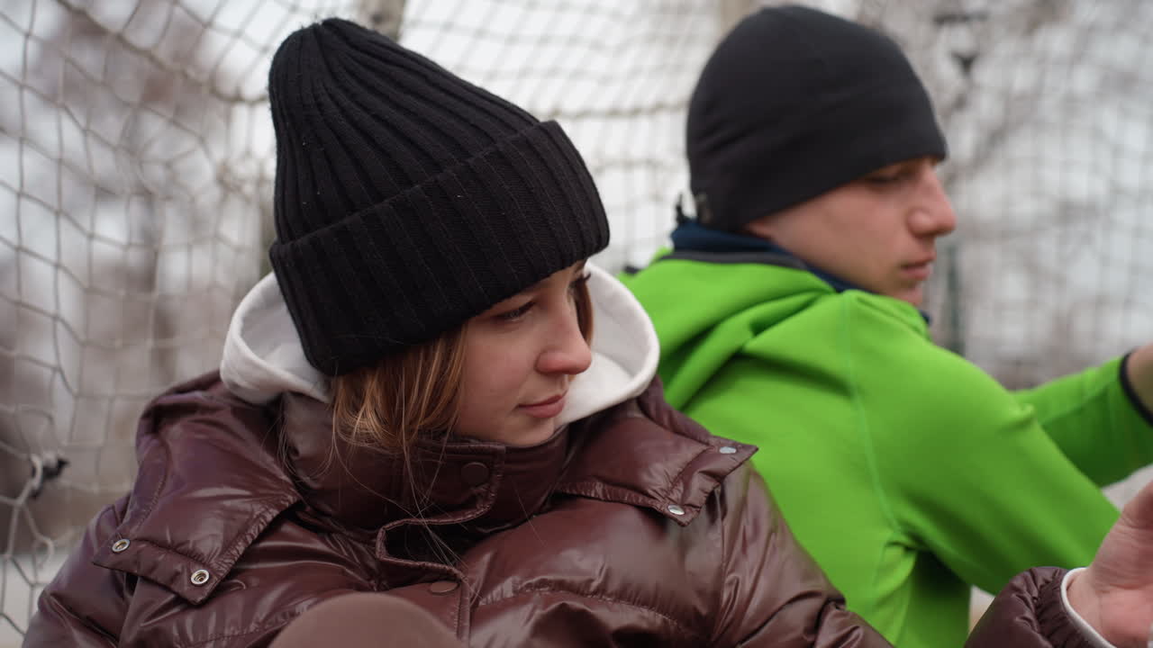 Задумчивый подросток-спортсмен, девушка-подросток в зимней куртке наблюдает за товарищем по команде во время перерыва на тренировке, юная спортсменка в шапке размышляет после тренировки возле спортивного инвентаря и боковой линии