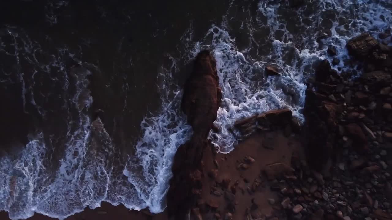Top view, drone hoovering a rock at the beach, water hitting rock during sunset