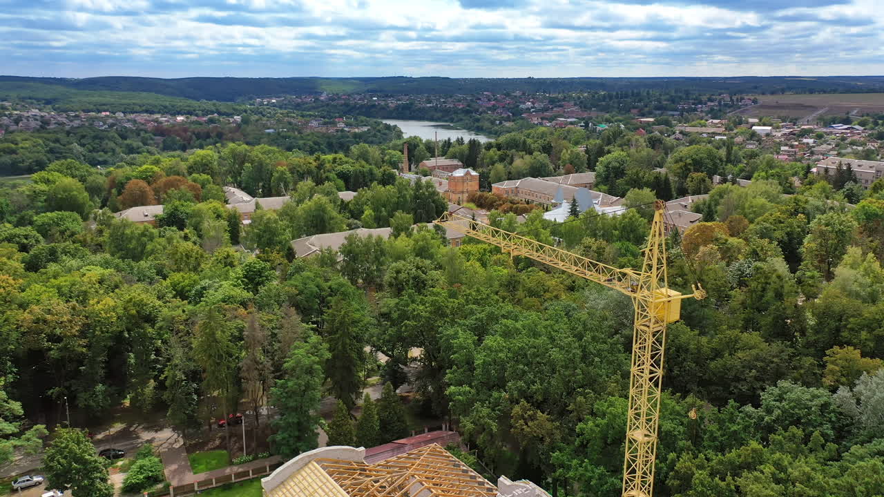 High crane works on building site with a house. Construction of a modern ecologic skyscraper with many green trees nearby.