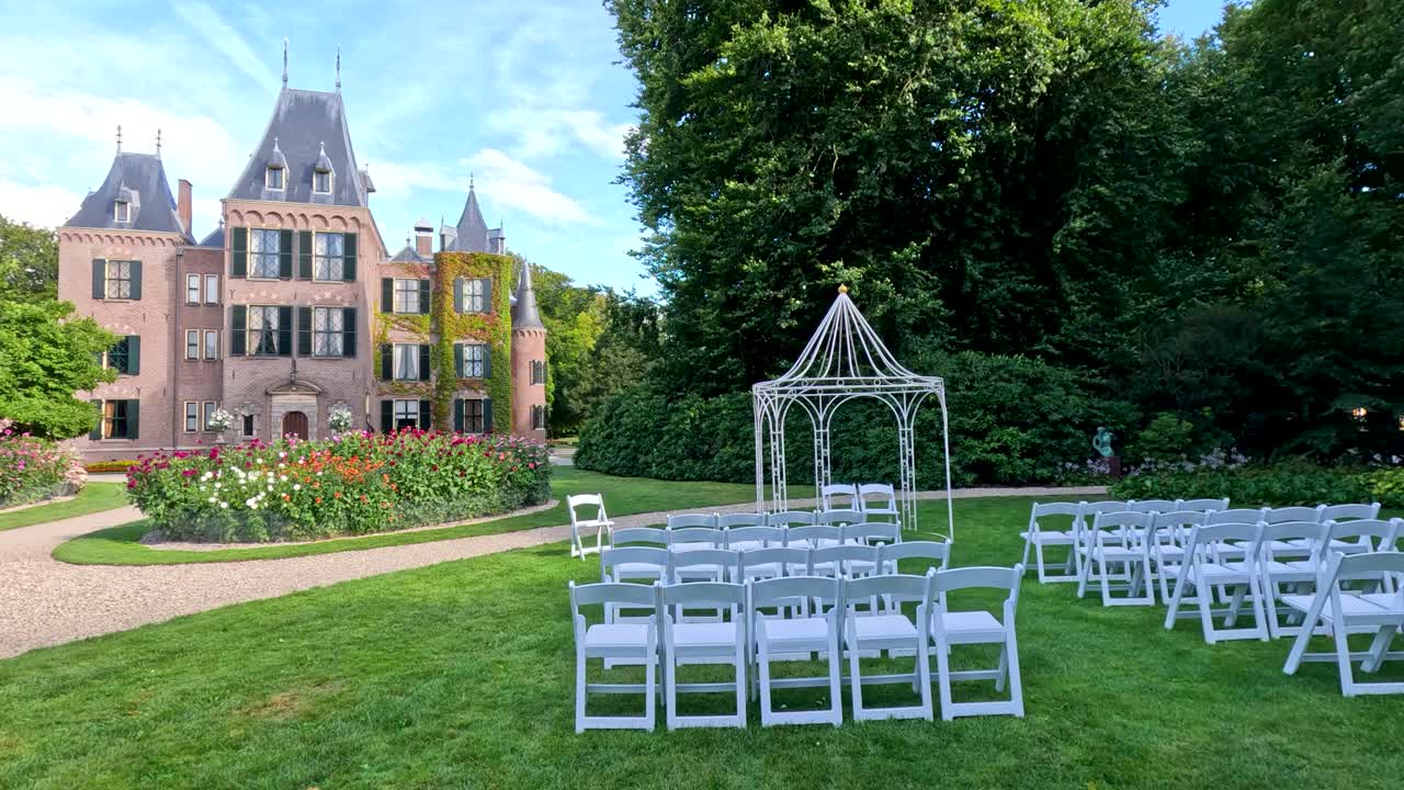 Rows of white chairs and a decorative arch are arranged on a manicured lawn for a wedding ceremony, with a castle and lush gardens in bright daylight. Subtle camera movement reveals the elegant outdoor setting