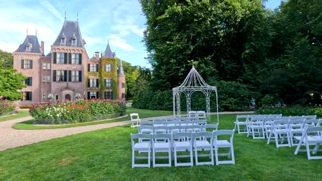 rows of white chairs and a decorative arch are arranged on a manicured lawn for a wedding ceremony, with a castle and lush gardens in bright daylight. subtle camera movement reveals the elegant outdoor setting