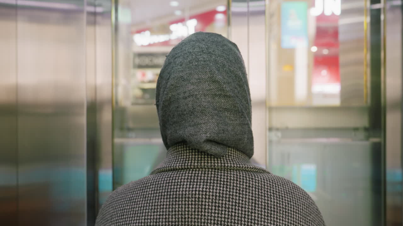 back view of woman wearing checkered coat and hood standing inside ascending elevator with reflective walls and blurred mall interior visible in background