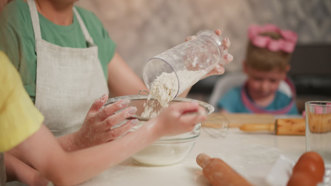 close up of child hands holding sieve while woman pours flour into bowl during baking session on white counter with eggs and rolling pin, while playful boy in background
