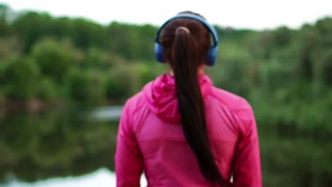 A-girl-in-a-pink-jacket-and-blue-headphones-stands-with-her-back-and-looks-at-the-river-early-in-the-morning-after-a-run