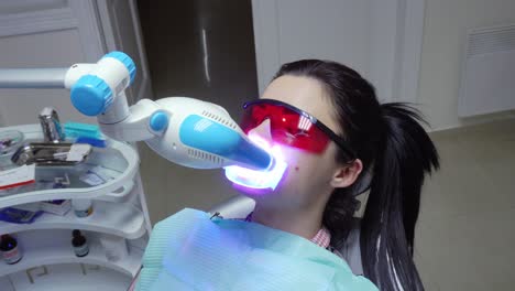 young woman with an expander in mouth and red protective glasses getting uv whitening. an ultra violet whitening machine in operation on a patient's teeth