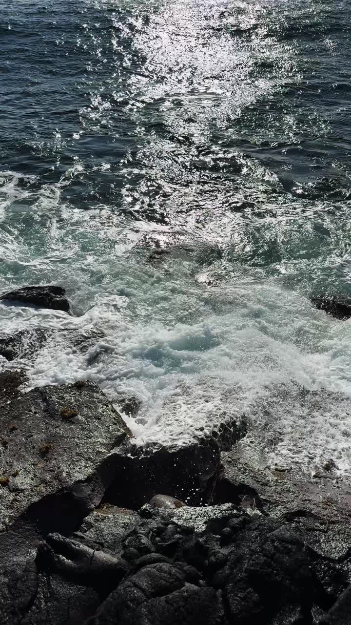 Vista dall'alto 4K verticale di onde oceaniche che si infrangono contro scure rocce vulcaniche, con la luce solare intensa che si riflette sulla superficie dell'acqua in movimento.