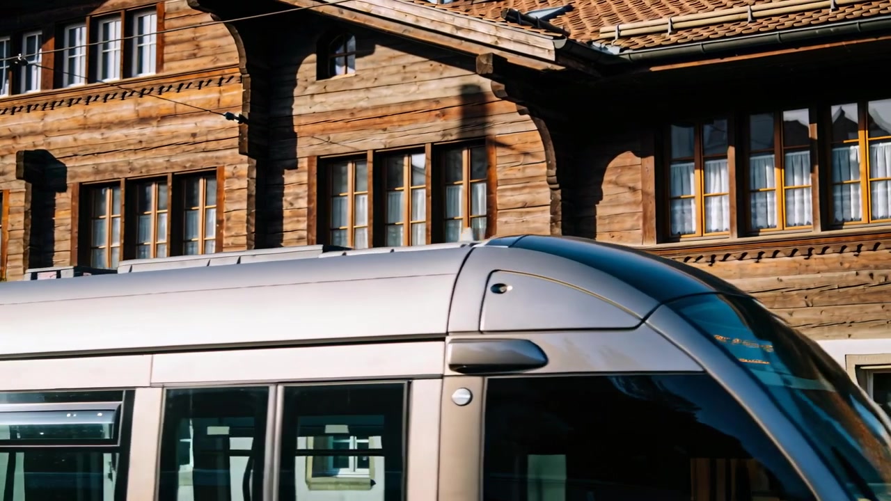 Moving articulated tram entering frame, crossing road past wood facade, pantograph on overhead wire
