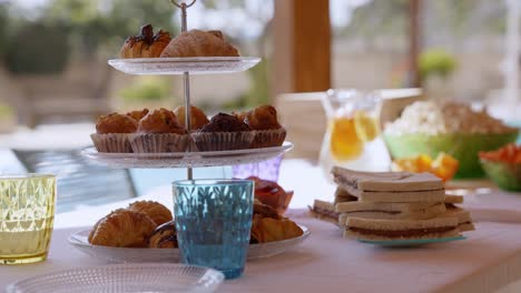 table with sweets for a poolside party in summer, with a tray with croissants, sandwich, popcorn, crudités, coloured glasses, transparent plates and white tablecloth